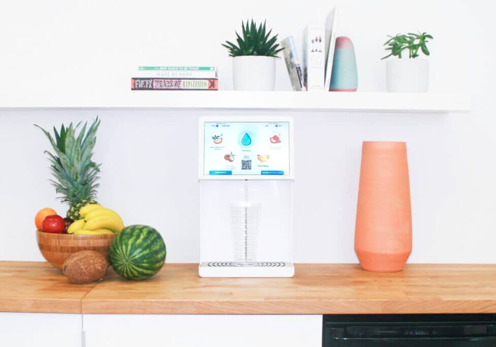 Bevi countertop water dispenser on office kitchen counter between fruit bowl and orange glass with small shelf above