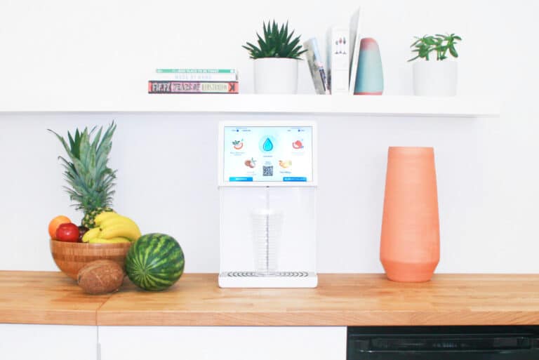Bevi countertop water dispenser on office kitchen counter between fruit bowl and orange glass with small shelf above