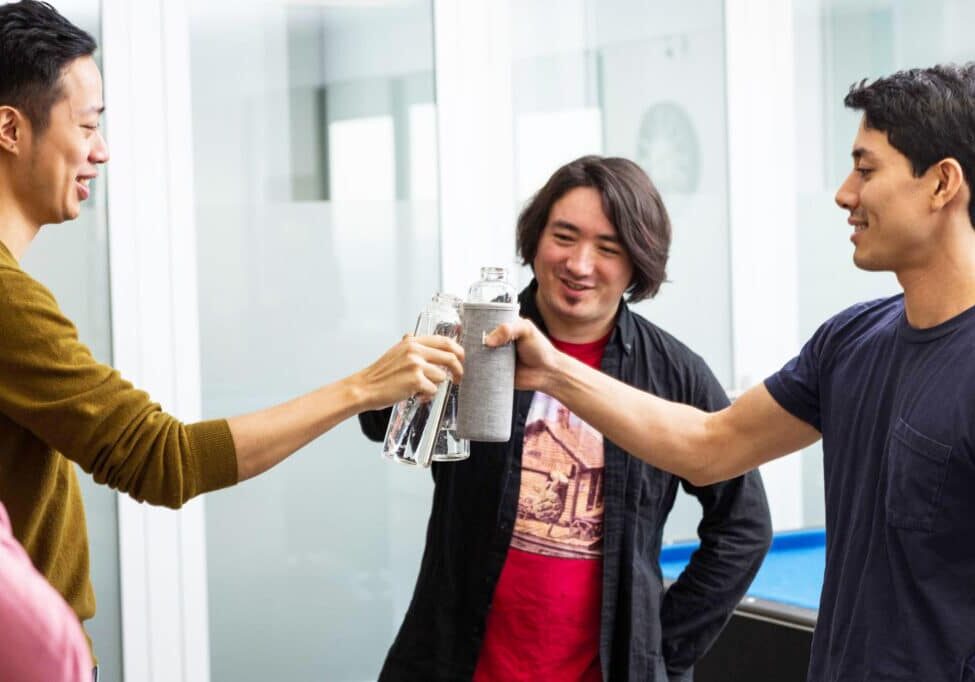 Three young men toasting with water bottles in an office kitchen