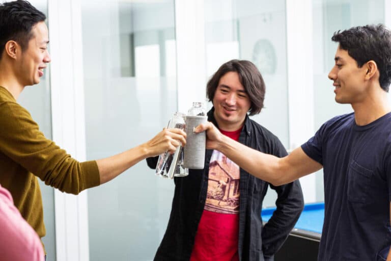 Three young men toasting with water bottles in an office kitchen