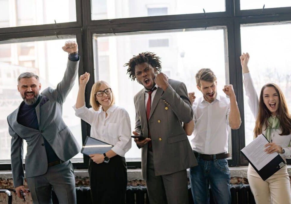 5 people in an office with smiles on their face and their hands raised cheering with happiness