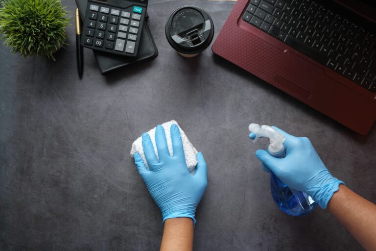 Hands with gloves, a towel, and cleaning spray wiping down a desk